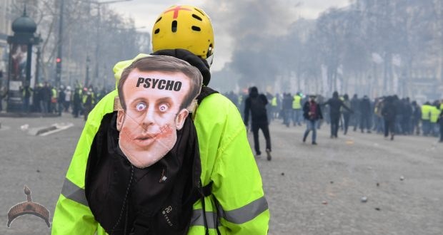 A Yellow Vest protester wearing a mask depicting the French President on which is written the word 'psycho' looks at fellow protesters in Paris on March 16, 2019, during the 18th consecutive Saturday of demonstrations called by the 'Yellow Vest' (gilets jaunes) movement. - Demonstrators hit French city streets again on March 16, for a 18th consecutive week of nationwide protest against the French President's policies and his top-down style of governing, high cost of living, government tax reforms and for more "social and economic justice." (Photo by Alain JOCARD / AFP)